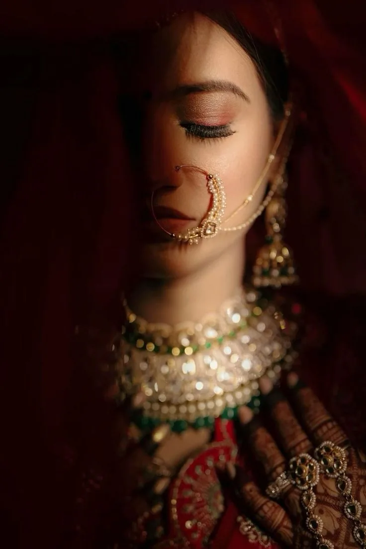 Bride under a red veil with her eyes lowered, the nath and kundan choker catching the light