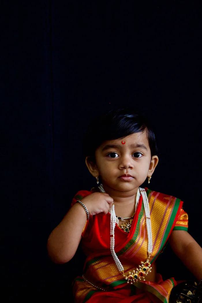 Child in a traditional red and gold outfit looking straight at the camera against a dark studio backdrop